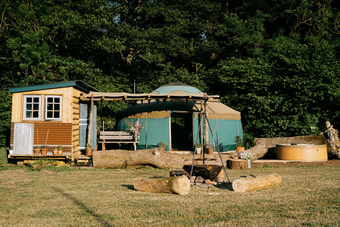 A charming glamping site at Southleigh Farm features a tiny wooden house, a large canvas tent, and a Meadow Yurt. A pergola with a bench and a fire pit with logs arranged around it enhance the space. Nearby, a hot tub is surrounded by lush greenery, creating a serene and inviting outdoor retreat.