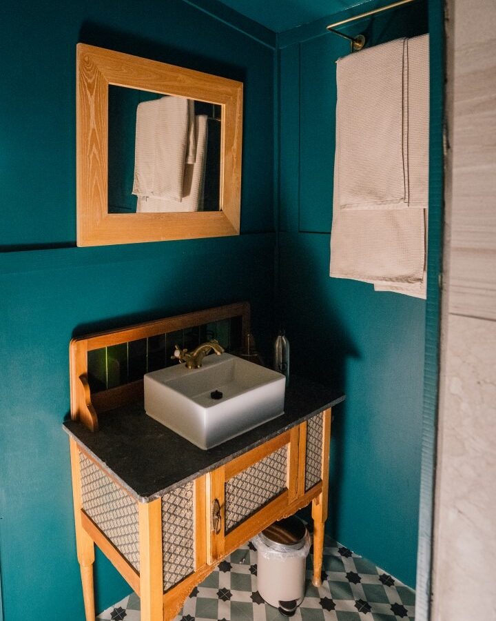 A small bathroom with teal walls and a matching ceiling, reminiscent of the serene atmosphere at Southleigh Farm, featuring a wooden vanity with a black countertop and a white sink. A mirror with a wooden frame hangs above the sink. Towels are neatly folded on a brass towel rack, and the floor has a geometric tile pattern.