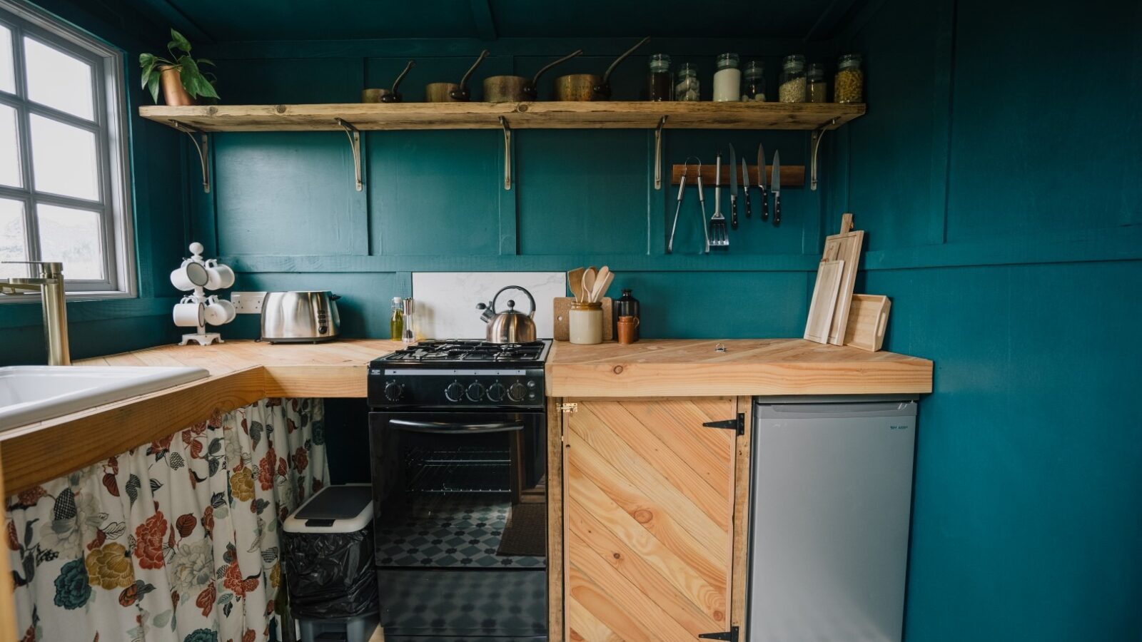 A compact kitchen at Southleigh Farm with teal walls and a wooden countertop. It features a small fridge, gas stove, and various utensils. A shelf holds jars and pots, while a patterned curtain covers storage under the sink. A window provides natural light.