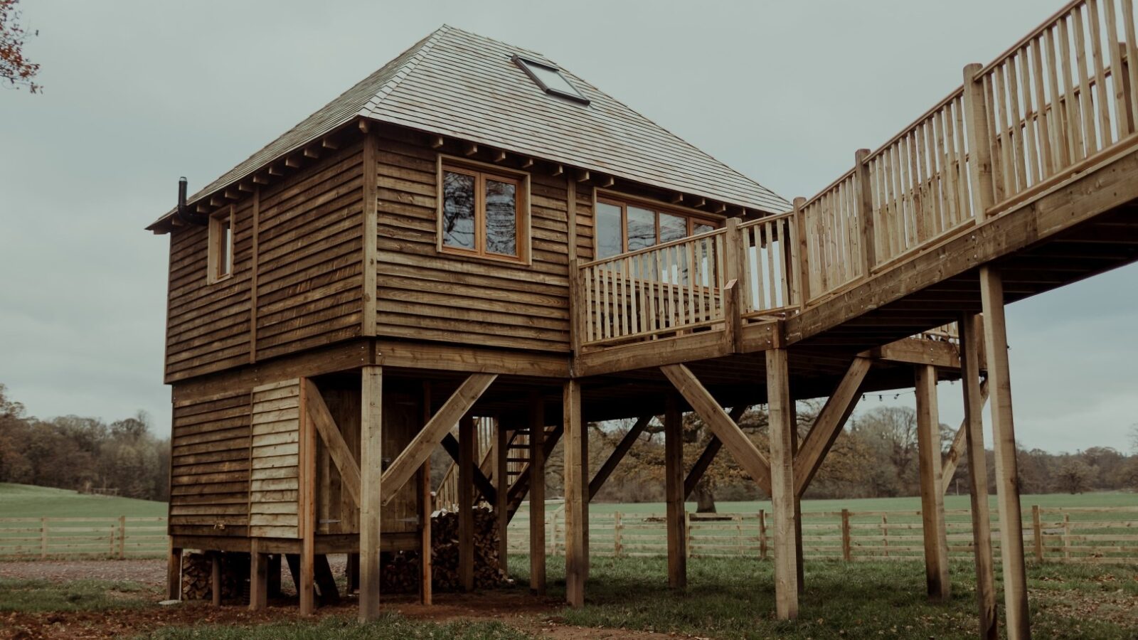 Nestled in a grassy field, the Netherby treehouse-style cabin stands elevated on stilts. It features a slanted roof with a skylight and a wraparound deck with railing, connected to a sloping wooden walkway. Leafless trees and a wooden fence frame the picturesque background.