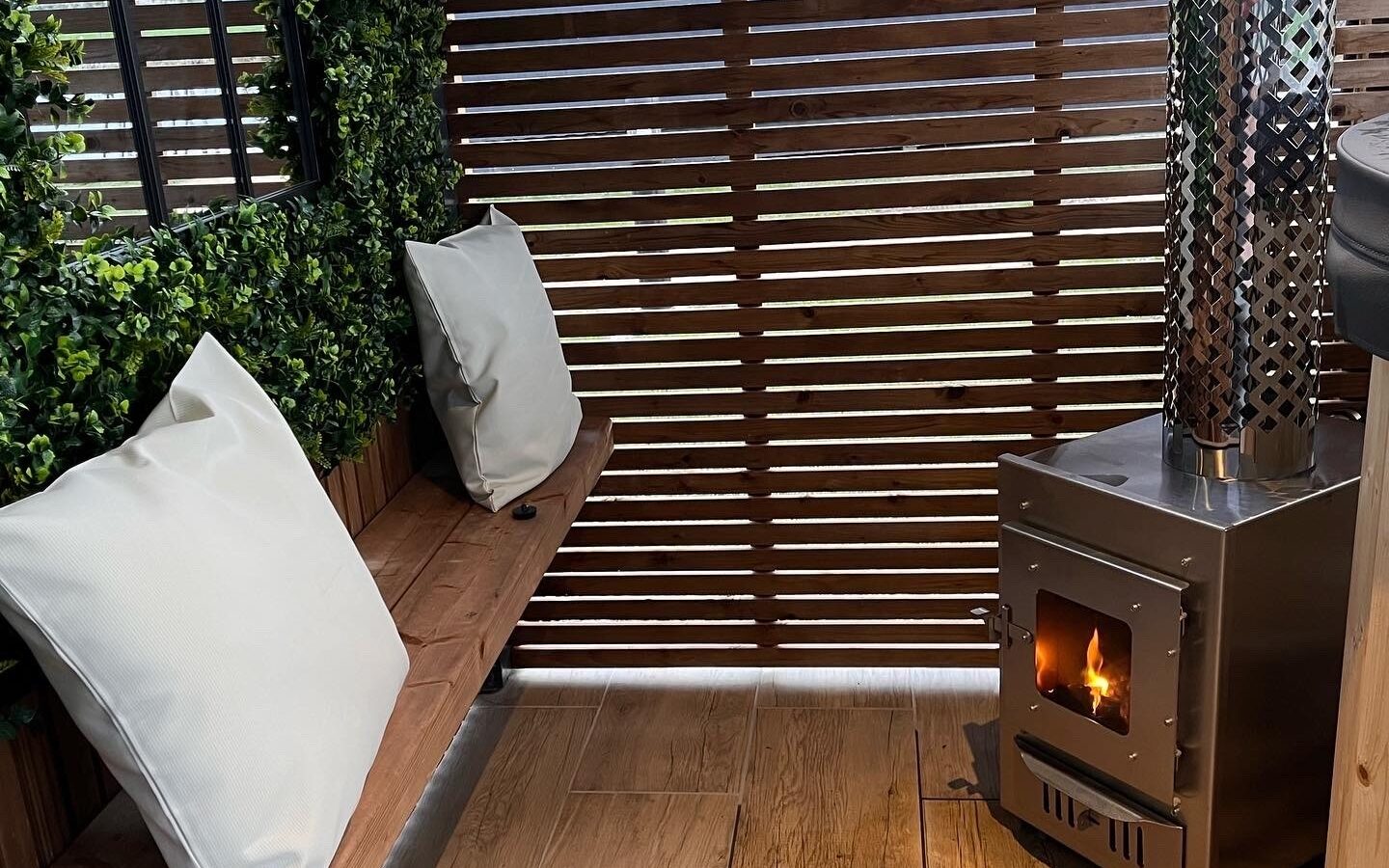 A cozy corner with a wooden bench and two white pillows is situated beside a small metal wood-burning stove with a visible fire. Enclosed by wooden slatted walls and decorated with lush greenery, this glamping spot on New Farm creates a warm and inviting ambiance.