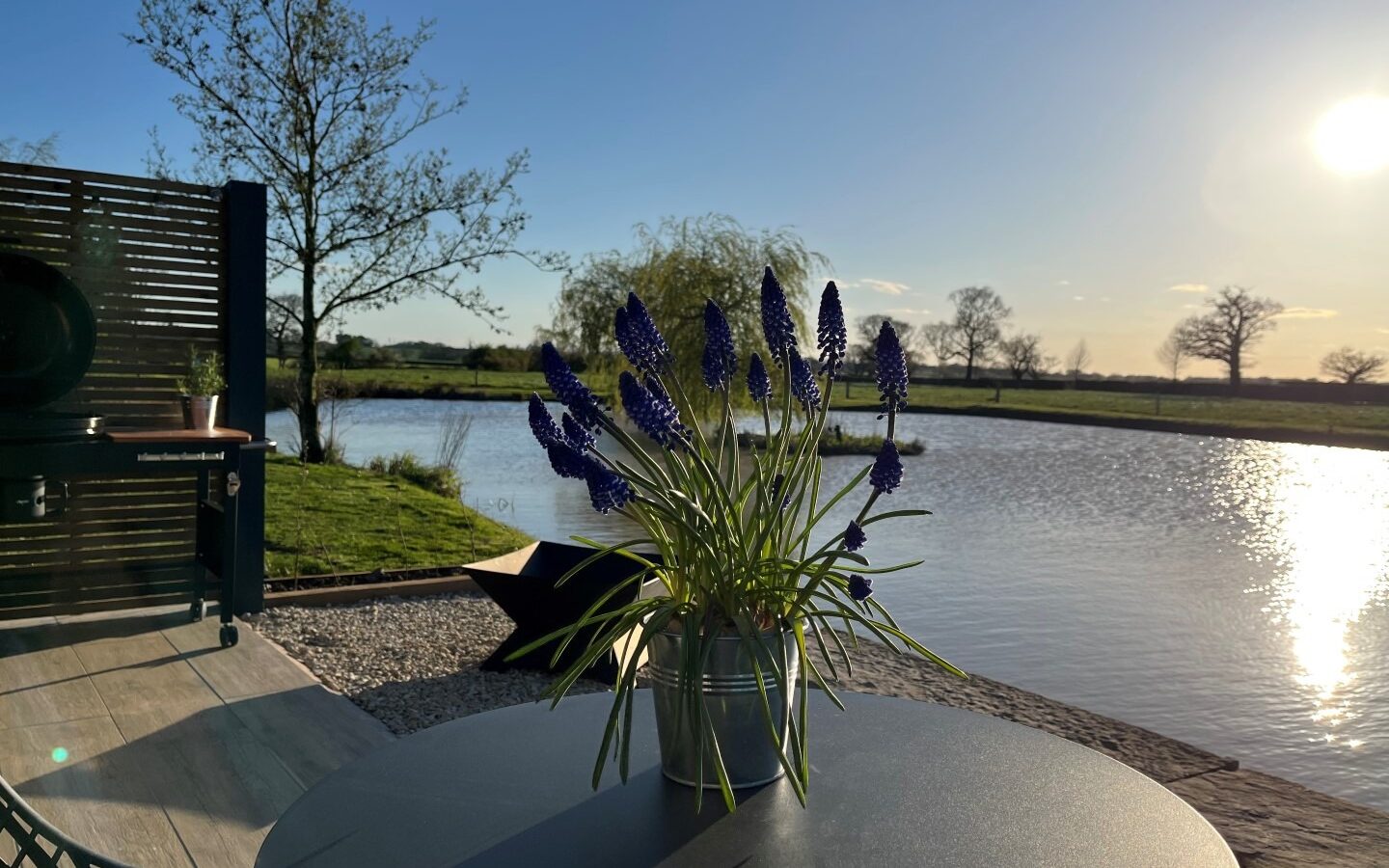 A bunch of purple flowers in a metal pot sits on a round table by a calm pond. The sun is low in the sky, casting a warm glow over the water and the surrounding trees. New Farm's glamping setup includes a grill and wooden fence visible on the patio area to the left.