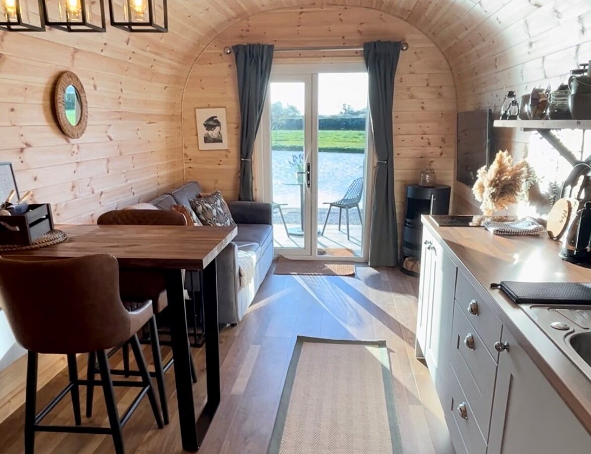 A cozy, wood-paneled tiny home interior featuring a living area with a brown sofa and a black table with two high chairs. The kitchenette includes white cabinets, a sink, and shelves. Large glass doors with curtains open to a view of a lake and greenery, perfect for your new glamping adventure.