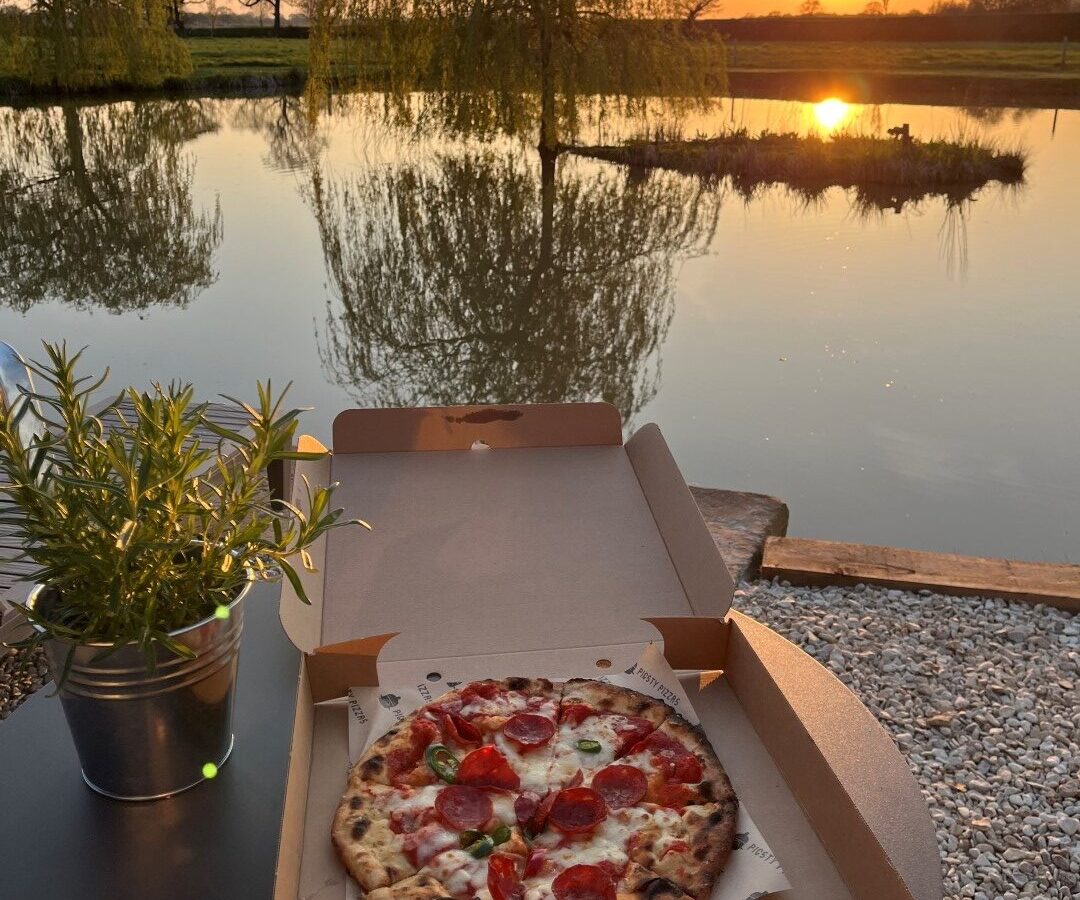 A pizza in an open box sits on a table next to a potted plant, overlooking a serene pond reflecting the sunset at New Farm. The sky is a warm orange and yellow, with trees and greenery framing the tranquil scene, evoking the peaceful ambiance of glamping.