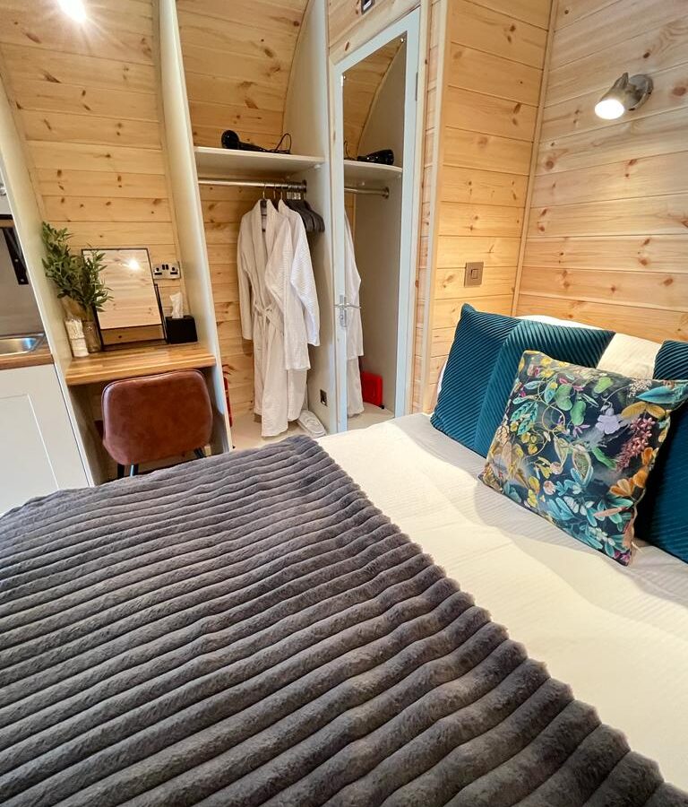 A cozy, modern bedroom with wood-paneled walls features a neatly made bed with a gray striped blanket and decorative pillows. To the left, a small desk with a brown chair sits beside an open closet with robes. Soft lighting adds to the warm atmosphere, perfect for anyone seeking an upscale glamping experience.