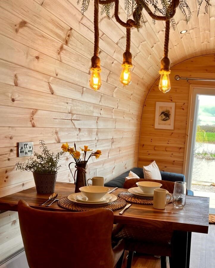 A cozy wooden cabin interior at New Farm Glamping features a small dining table set for two with round placemats, bowls, mugs, and a vase of yellow flowers. Above the table, a rustic chandelier with three hanging Edison bulbs adds warm lighting. A window reveals a scenic outdoor farm view.