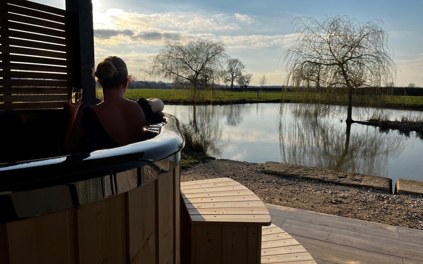 A person with tied-back hair relaxes in a wooden hot tub at New Farm, overlooking a serene lake with bare trees silhouetted against a setting sun. Wooden steps lead up to the tub, and the scene is framed by clear skies and calm water, perfect for glamping enthusiasts.