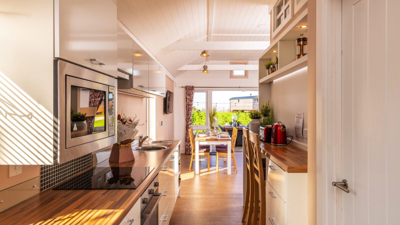 A bright, modern kitchen with wooden countertops, white cabinets, and stainless steel appliances. An open door at the end leads to a sunny dining area with a table set for two, overlooking a green garden. Several potted plants decorate the space, adding a natural touch reminiscent of rural retreats like Ockeridge.