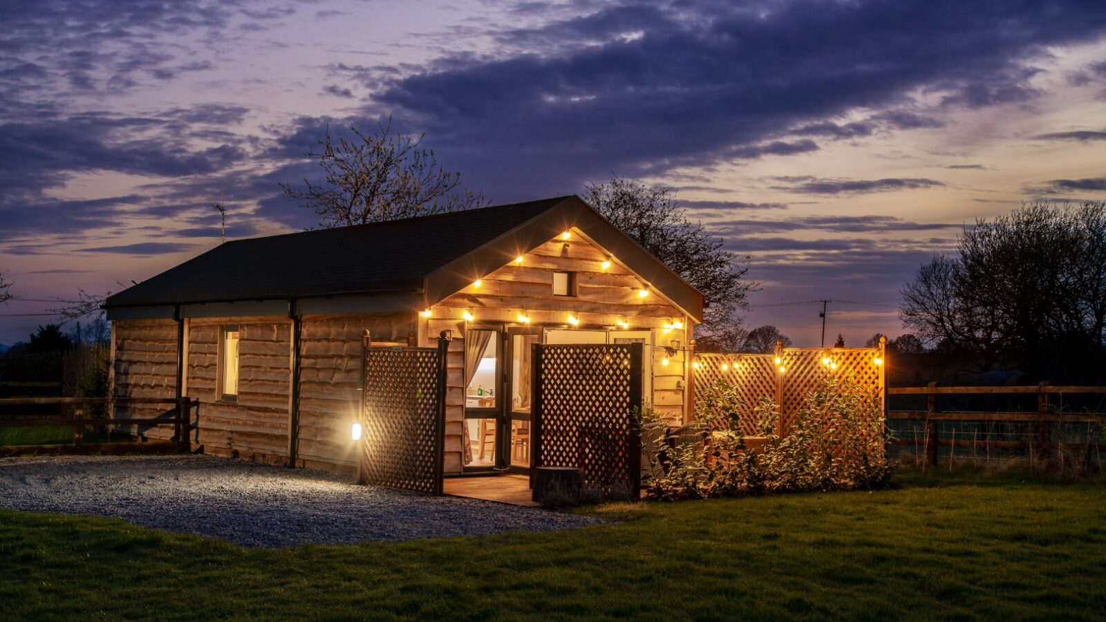 A cozy wooden cabin with warm string lights adorning the exterior sits against a twilight sky in the rural Ockeridge countryside. The windows glow invitingly, and a small patio is enclosed with a trellis fence. Trees and a cloudy purple sky provide a serene backdrop for this tranquil retreat.