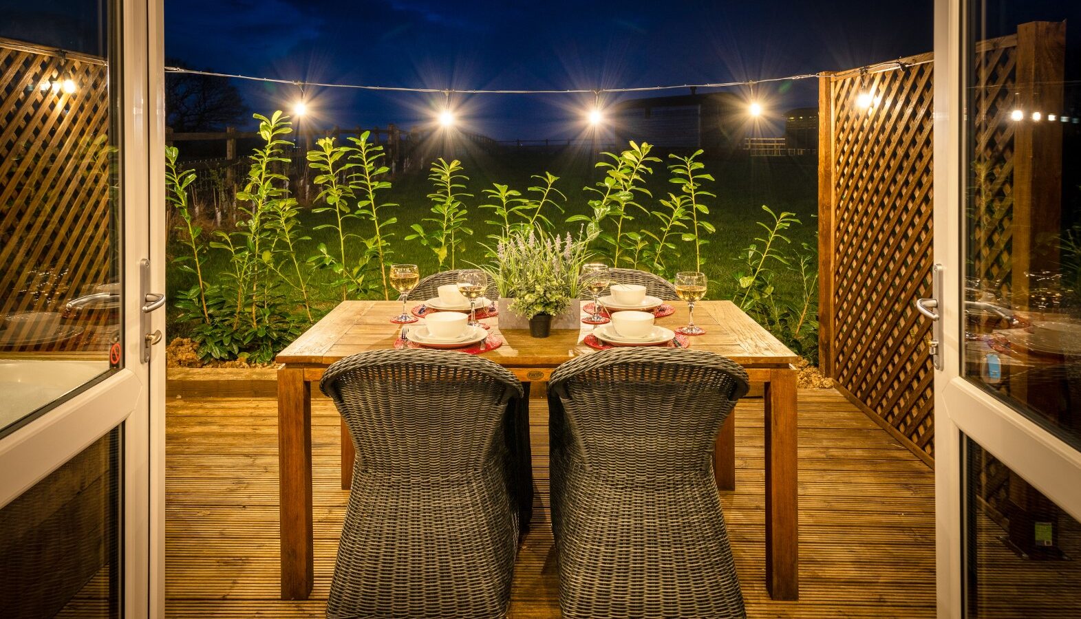 A cozy outdoor dining setup at night, framed by open doors. The wooden table is set with white plates and glasses, surrounded by four wicker chairs. String lights hang above, illuminating the scene. Tall green plants line the wooden lattice fence in the background, creating a perfect rural retreat at Ockeridge.