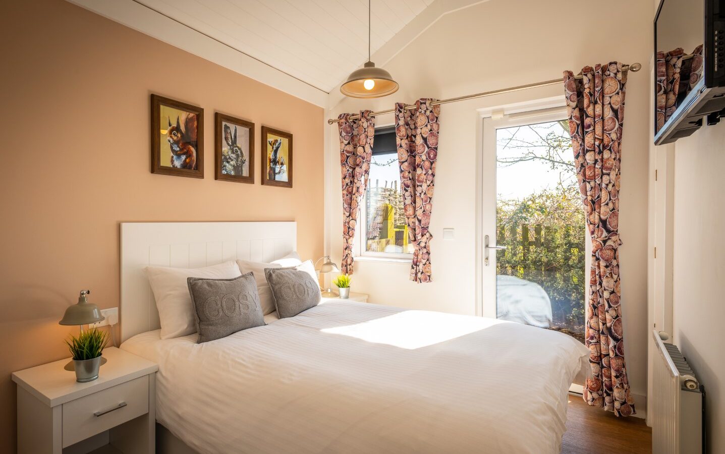 A cozy bedroom at Ockeridge featuring a large window and a glass door that let in natural light. The room offers a white bed with two gray pillows, bedside tables with lamps, floral curtains, three paintings on a pastel peach wall, and a small potted plant on a table—a perfect rural retreat.