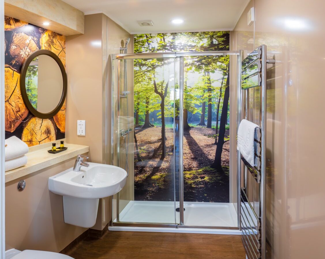 A modern bathroom with a large shower that has a glass door featuring a wooded forest scene. There's a white sink and round mirror on the left with towels and toiletries nearby. The walls are beige, and it has wooden flooring reminiscent of Rural Retreats. An electric towel heater is on the right.