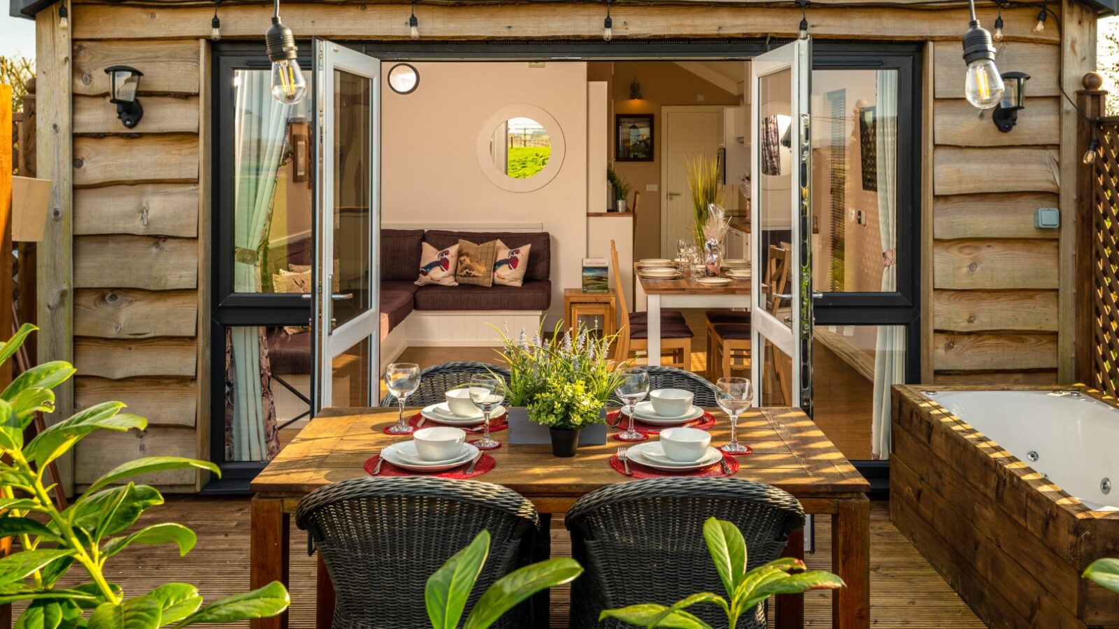 A cozy outdoor dining area with wicker chairs and a wooden table set for four. The table is adorned with a potted plant centerpiece. Double doors open into a warmly lit, rustic interior featuring a couch and a dining table. Greenery surrounds this rural retreat, creating an inviting Ockeridge ambiance.