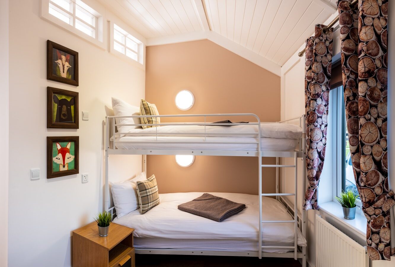 A cozy, compact bedroom at Ockeridge Retreats features a white metal bunk bed with crisp white linens and patterned pillows. The room has a warm peach accent wall, circular wall lights, wood flooring, and a bedside table with a small plant. Three animal-themed paintings adorn the wall.