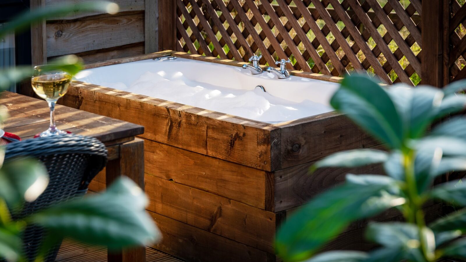 A luxurious outdoor bathtub with a wooden frame is filled with bubbles, set on a wooden deck beside a lattice privacy screen. A glass of white wine sits on a table nearby, partially obscured by lush green plants in the foreground—an idyllic retreat at Ockeridge Rural.