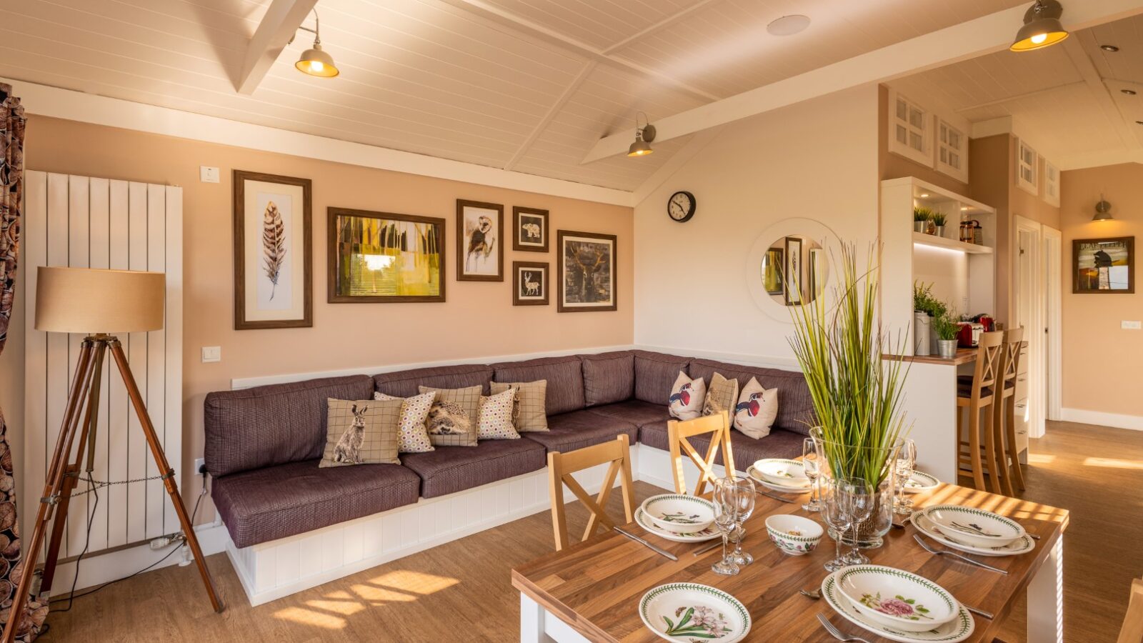 A cozy dining and living area with wooden floors and warm lighting evokes the charm of rural retreats. The Ockeridge-style dining table is set with plates and cutlery, while the living space has a cushioned, L-shaped bench with decorative pillows. Wall art and potted plants add a homey touch to the space.