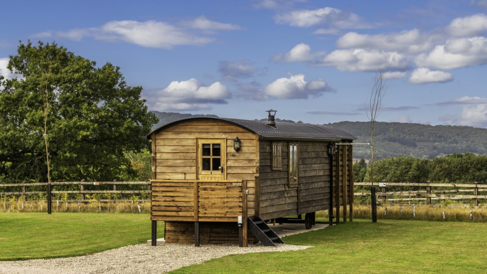 A small wooden cabin on stilts, with a front porch and steps, is set on a grassy landscape. A gravel pathway leads to the Ockeridge cabin. Trees and rolling hills are visible in the background under a partly cloudy blue sky, making it one of the perfect rural retreats.