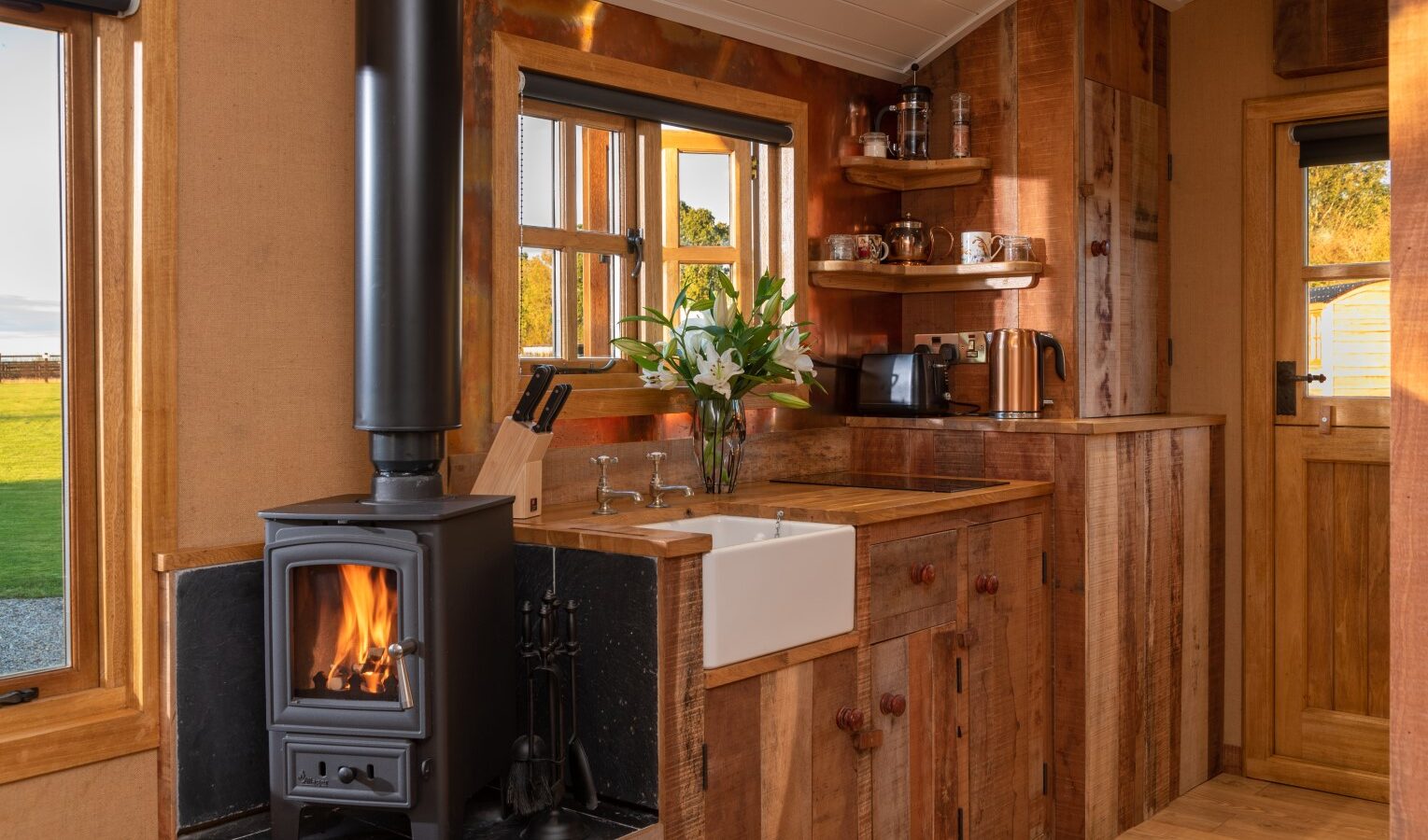 A cozy rustic kitchen from Ockeridge features a wooden interior with a black wood-burning stove on the left, a white farmhouse sink, wooden cabinets, and shelves with kitchenware. The window and door offer a view of greenery outside. A vase with white flowers sits by the sink in this charming Rural Retreat.