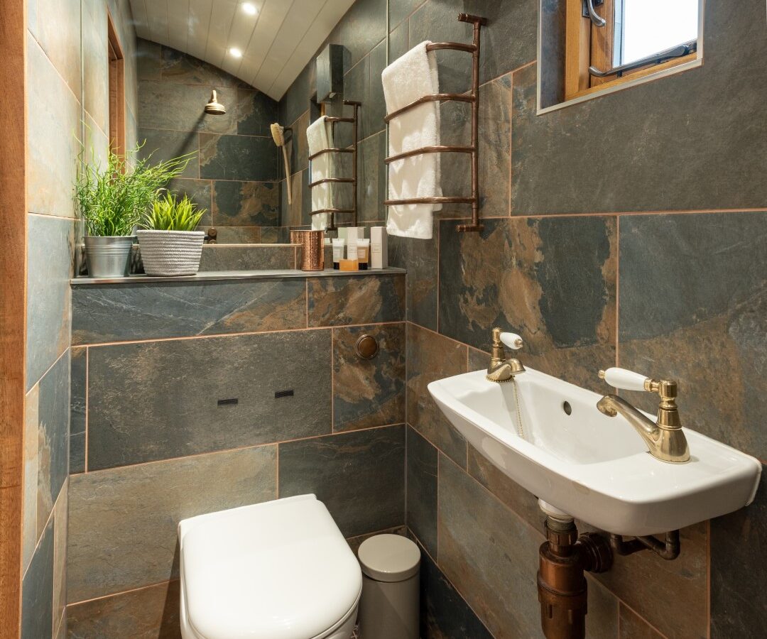 A modern bathroom with stone-tiled walls and floor, reminiscent of the elegance found at Ockeridge Rural Retreats. It features a wall-mounted toilet, a small white sink with brass fixtures, a rectangular mirror, and a towel rack with white towels. A window provides natural light, complemented by decorative potted plants.