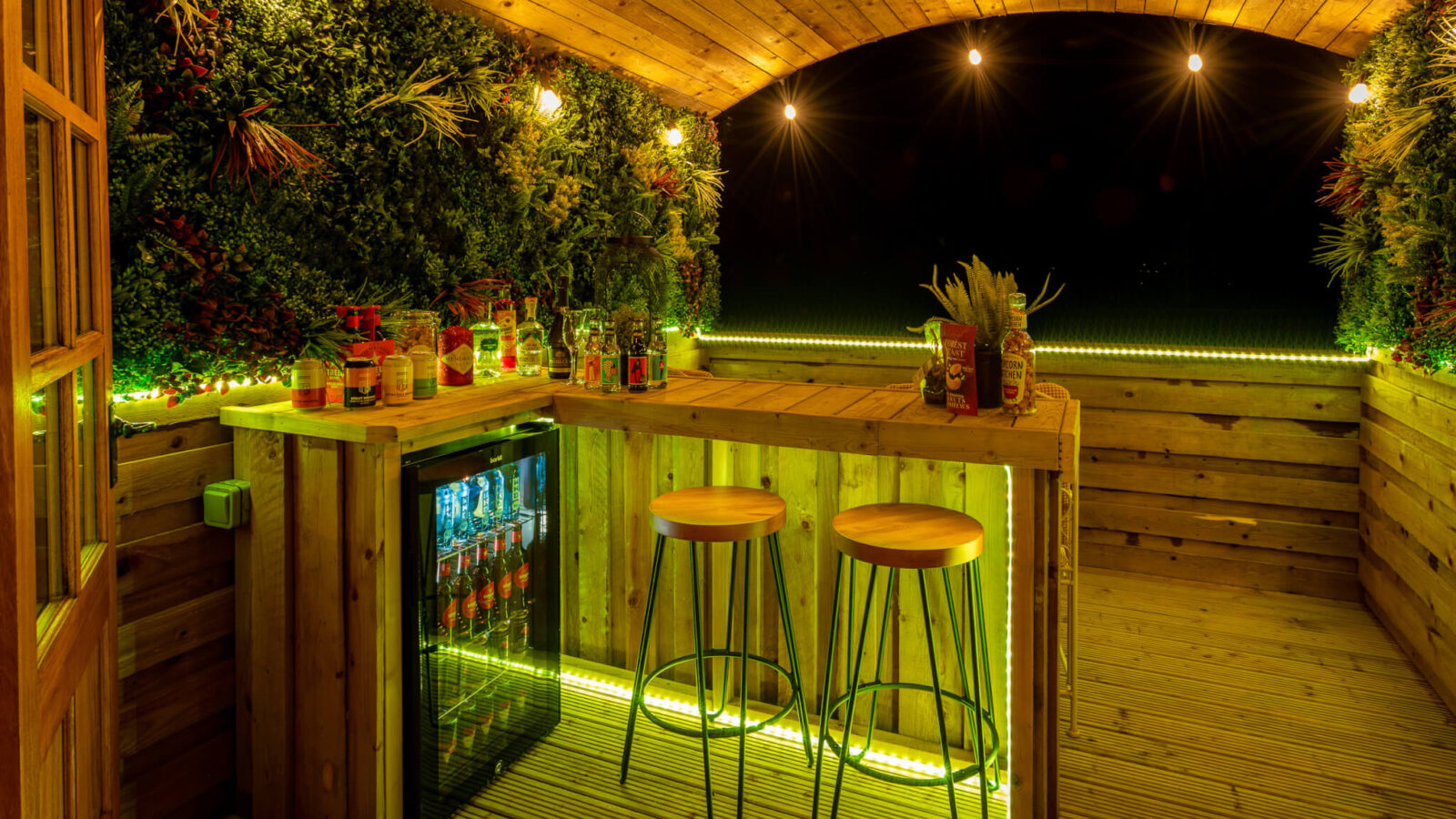 A cozy wooden bar at Ockeridge Rural Retreats features a green foliage backdrop, illuminated by warm string lights and green LED lights. Two wooden stools are placed at the bar, which is stocked with various drinks and a mini-fridge underneath. The ceiling is arched, adding to the rustic charm.