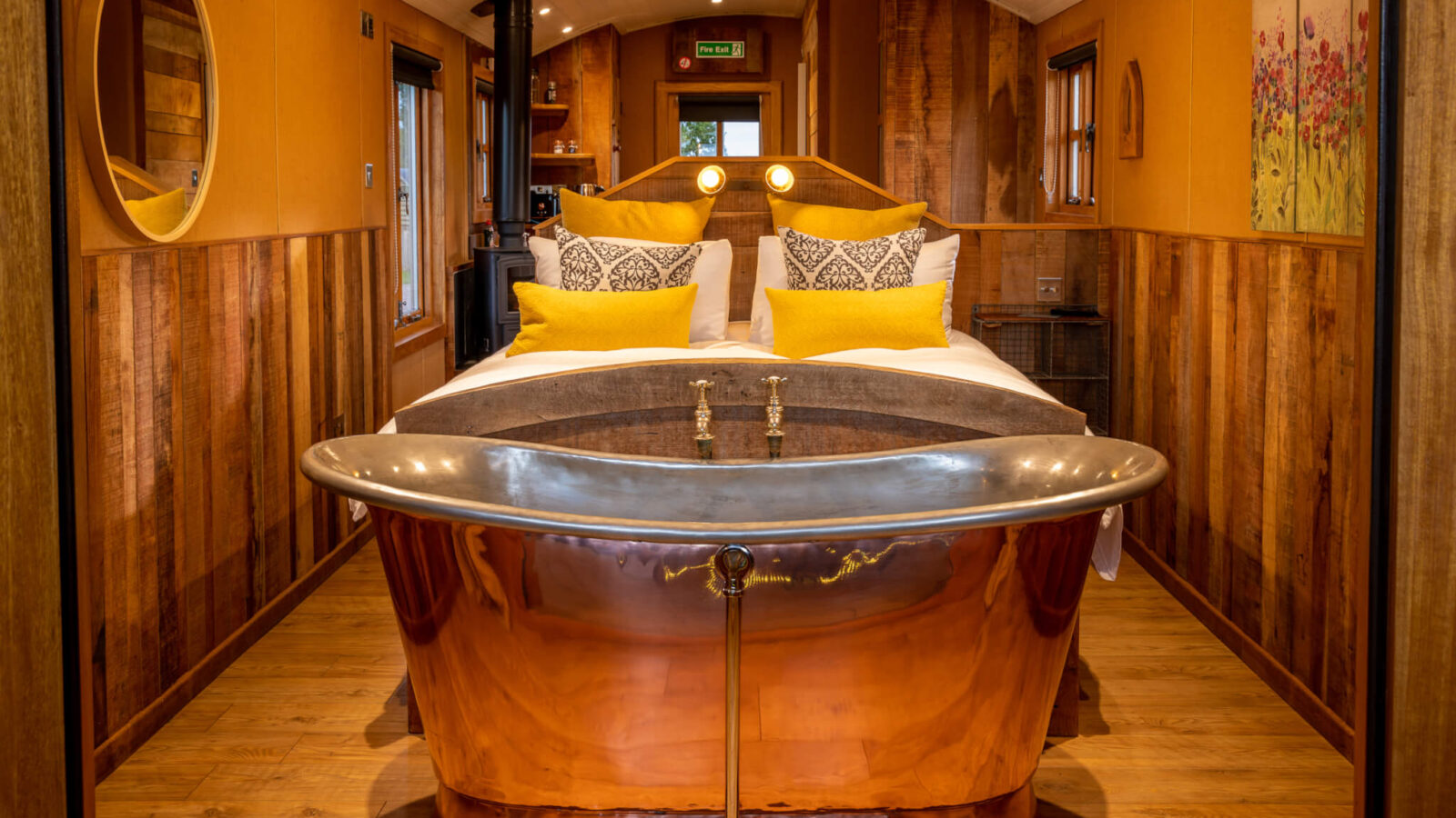 A cozy bedroom retreat features a large copper bathtub at the foot of a neatly made bed with yellow pillows. Warmly lit with wooden paneling on the walls, this rural haven is adorned with various decorative items, including wall art and mirrors, creating a rustic ambiance.