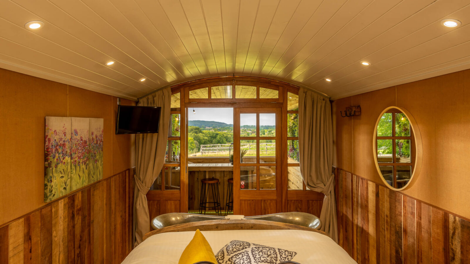 A cozy room with a wooden interior, featuring a bed with patterned pillows and a mustard throw pillow. The room, perfect for rural retreats in Ockeridge, has a large window with curtains, a landscape painting on the left wall, a round mirror on the right, a small TV, and a bar stool by the window.