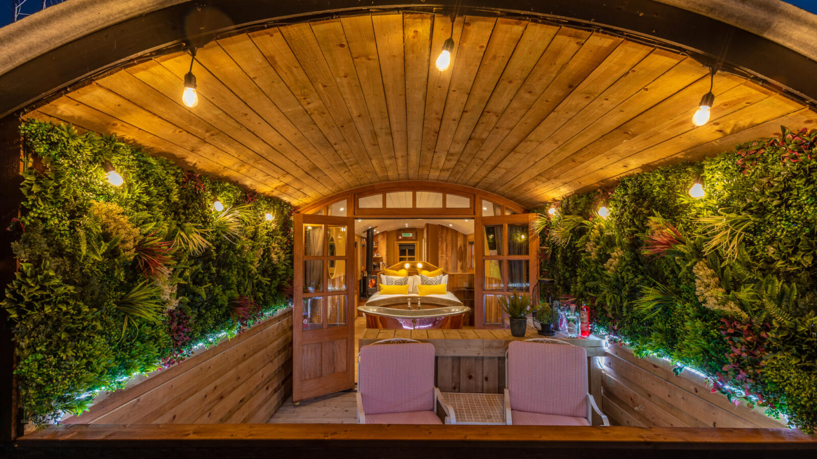 A cozy, rustic cabin terrace at night, with string lights illuminating a wooden ceiling and lush, green plant decorations on the walls. Two pink chairs are placed at the entrance, leading into a warmly lit interior with a bed and a small table—a perfect spot for Ockeridge Rural Retreats.