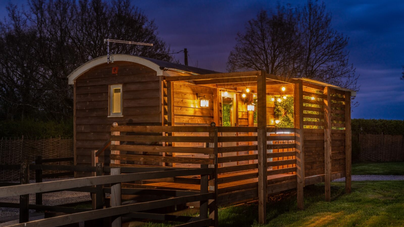 A cozy wooden cabin at Ockeridge Rural Retreats, illuminated by warm lights, stands in a grassy area during dusk. The cabin has a small porch with a wooden railing and is surrounded by bare trees and a dark blue sky. The ambient light creates a welcoming atmosphere.