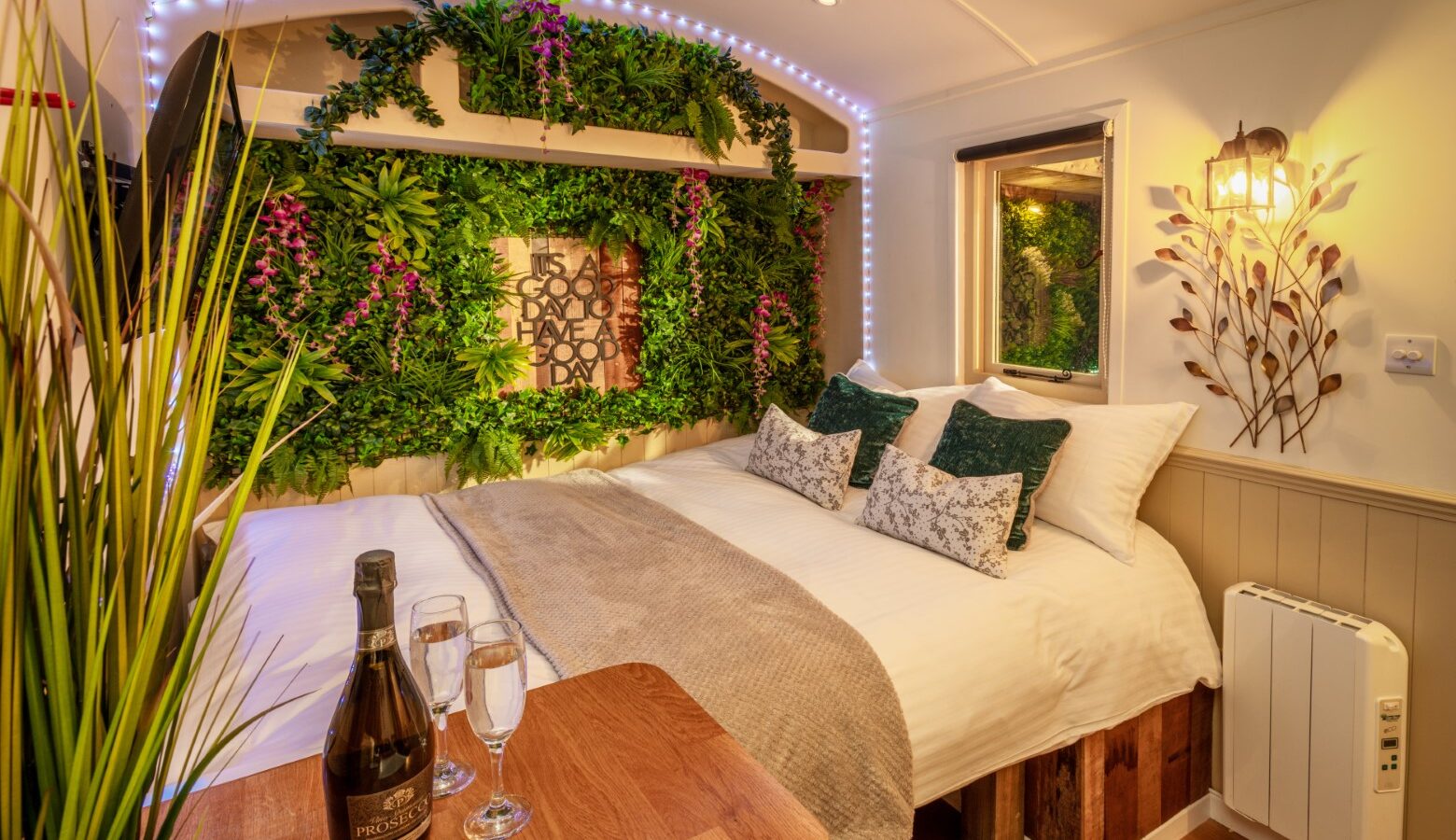 A cozy, elegant bedroom at Ockeridge Retreats features a bed adorned with white linens and green accent pillows. Above the headboard, a lush green plant wall with purple flowers adds charm. A bottle of wine and two glasses sit on a small wooden table, bathed in warm lighting that enhances this rural haven's ambiance.