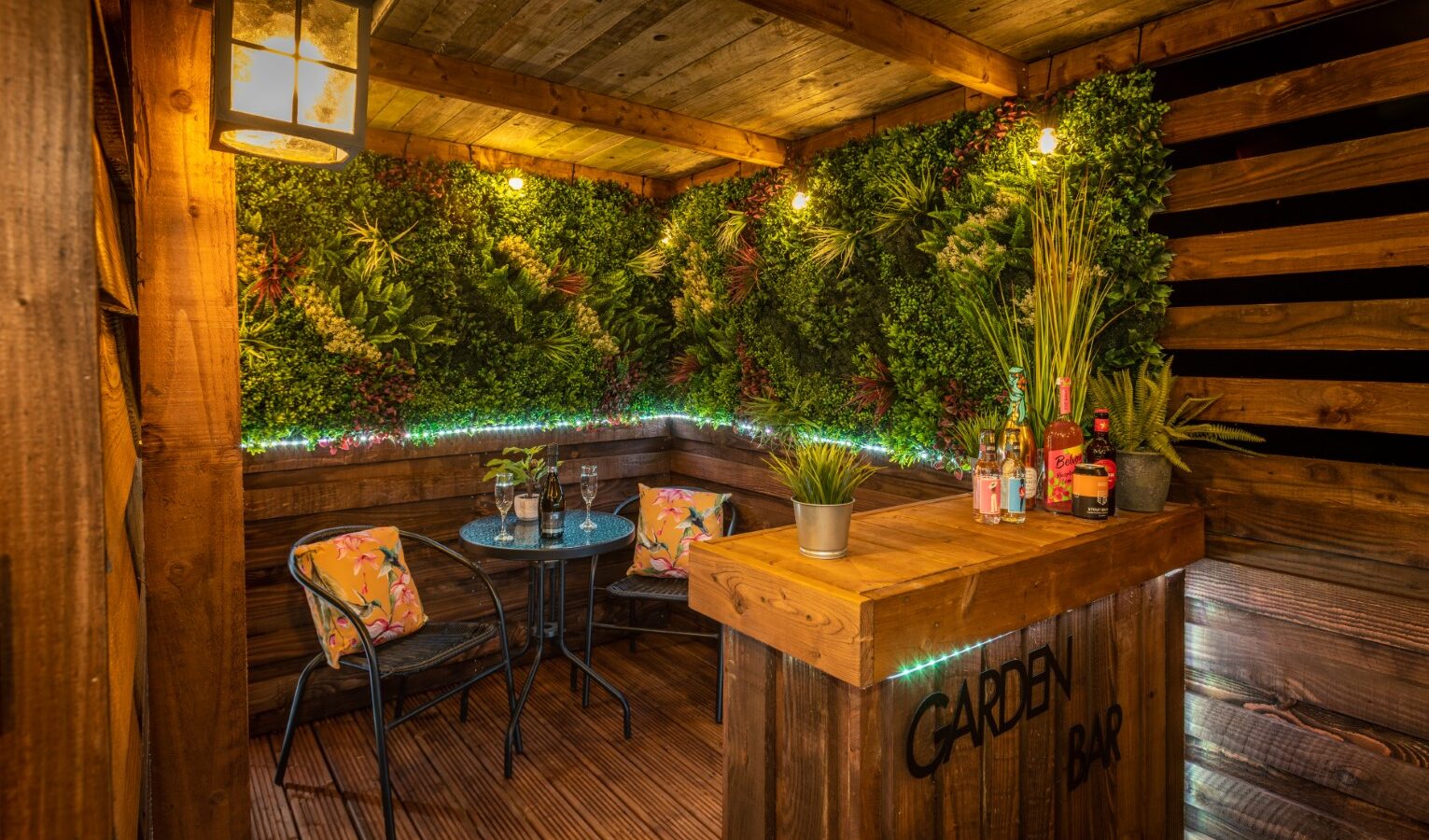 A cozy garden bar with wooden walls and flooring, decorated with lush green plants and string lights. The bar counter has various bottles and a plant. There's a small table and two chairs with floral cushions. A lantern light is mounted on the wall, adding warmth to this charming Ockeridge rural retreat.