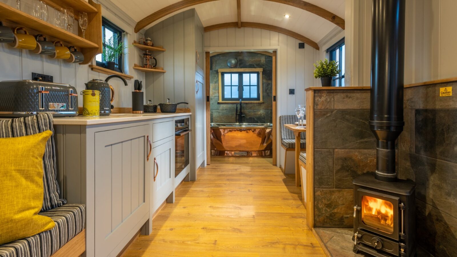 A cozy and stylishly designed tiny home interior at Ockeridge Retreats features an arched wooden ceiling, a small wood-burning stove, a kitchen with white cabinetry, open wooden shelves, and a built-in bench with cushions. In the background is a rustic bathroom with a copper tub that enhances its rural charm.