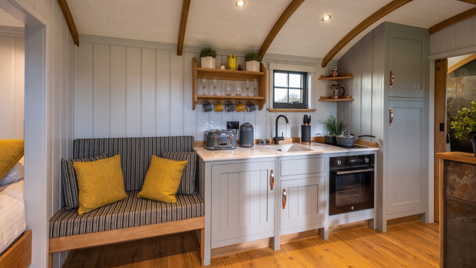 A cozy, well-lit kitchen in a small Ockeridge retreat featuring wooden beams and flooring. There is a bench with striped cushions, a countertop with appliances including a toaster and kettle, open shelving with glasses, and cabinets for storage. A small window allows natural light to flood this rural haven.