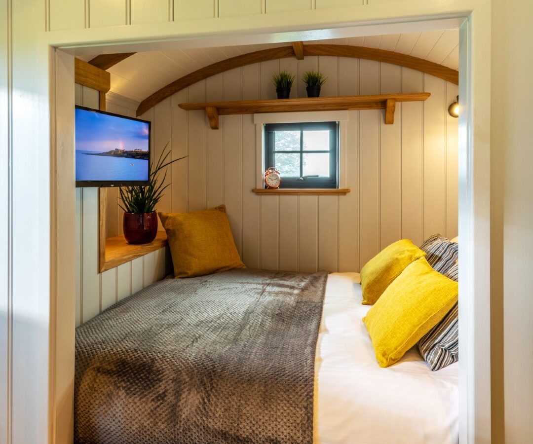 A cozy bedroom at Ockeridge Rural Retreats with a small window. The bed has a wooden frame, gray bedding, and yellow pillows. Shelves above the bed hold small plants, and a flat-screen TV is mounted on the left wall. The room has white paneled walls and a wooden ceiling.