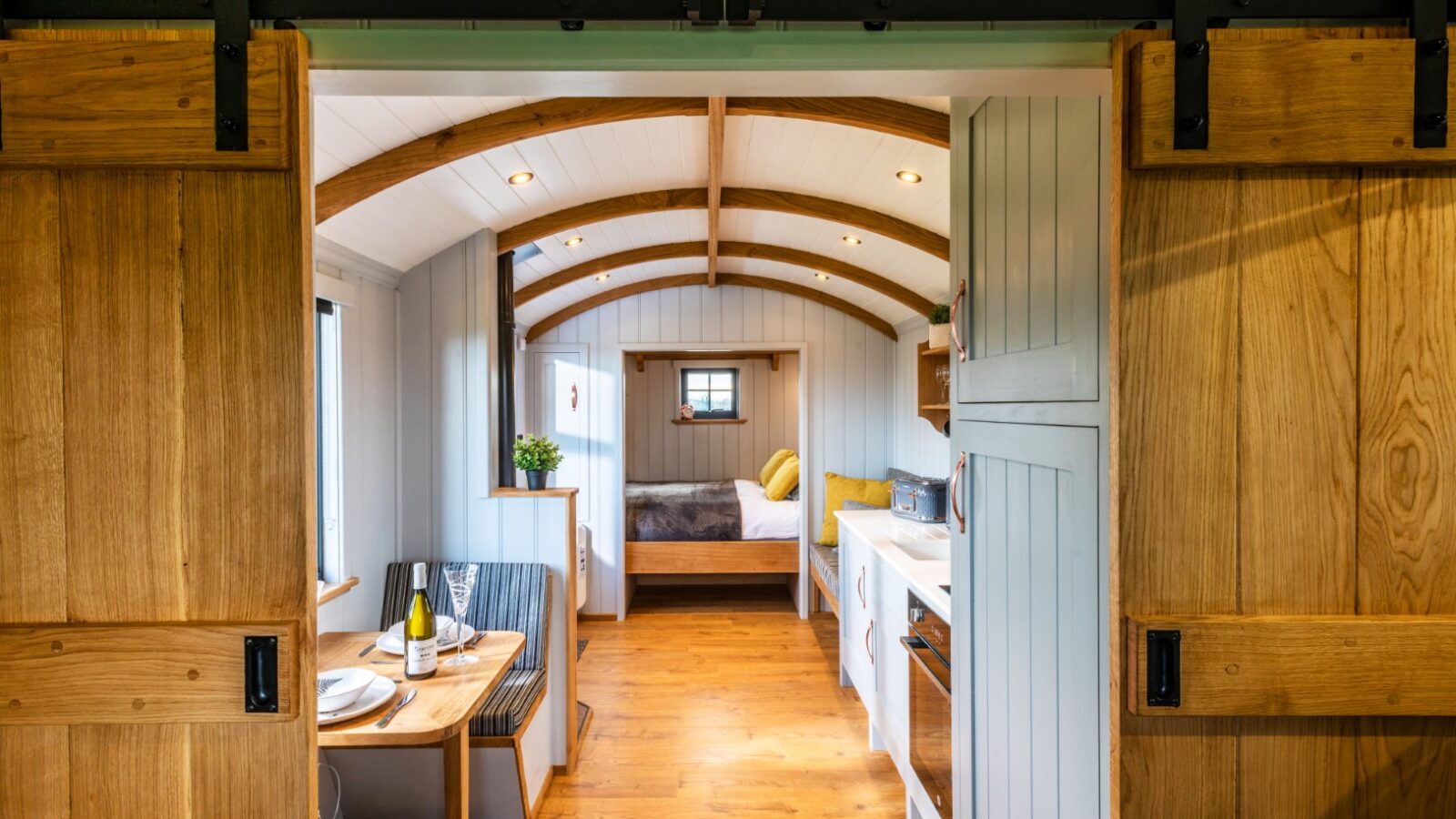 A cozy, well-lit tiny home interior at Ockeridge Rural Retreats features wooden sliding barn doors, a small dining area set for two, a compact kitchen with white cabinets, and a bed with white and yellow bedding at the far end. The ceiling boasts charming curved wooden beams.