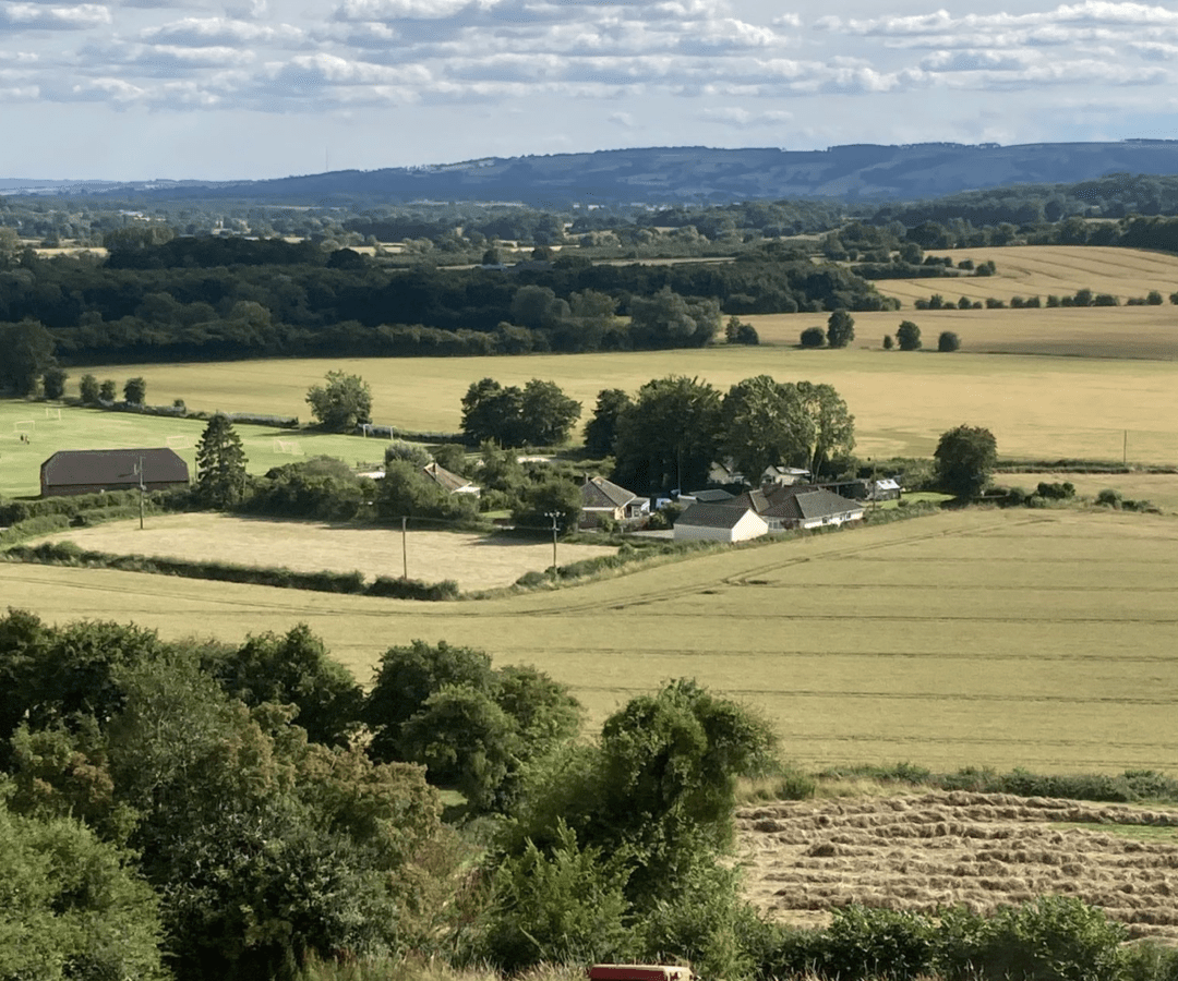 A tractor works in a field harvesting hay, with several hay bales scattered around. Near One Acre Wood, the field is encircled by trees and homes, with rolling hills and a cloudy sky in the background.