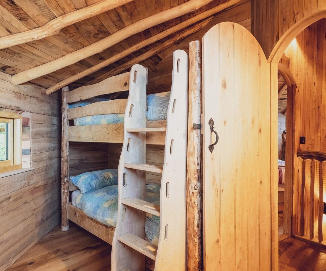 A cozy, rustic wooden bedroom in One Acre Wood features charming bunk beds standing under a slanted ceiling. The beds have a built-in wooden ladder and are adorned with blue bedding. A partially open wooden door is on the right, while a colorful rug adds warmth to the left side of the room.