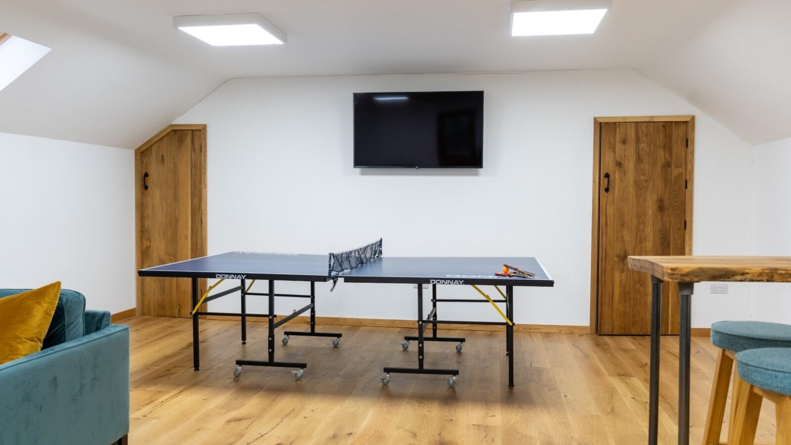 A game room with a ping pong table in the center, reminiscent of the rustic charm found in One Acre Wood. The room features wooden floors, a flat-screen TV mounted on the wall, and wooden doors on either side. A green couch with yellow cushions sits to the left, while a high wooden table with stools is to the right.