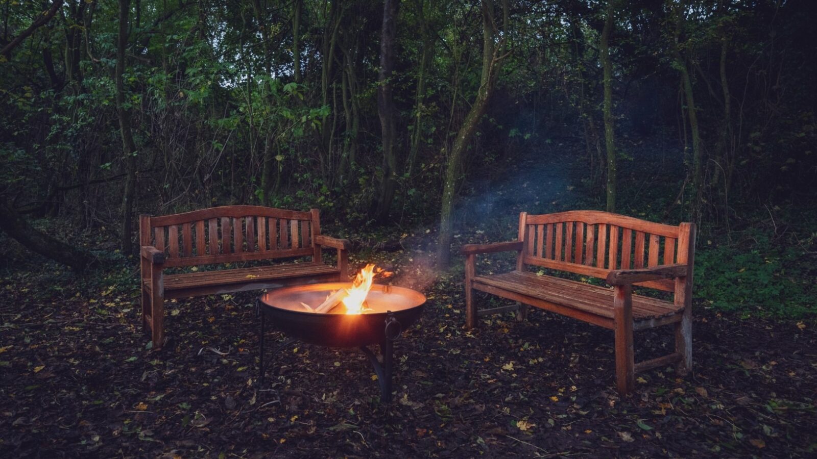 Two wooden benches face each other with a circular fire pit in the center. Situated in One Acre Wood, the benches are surrounded by tall trees. The fire pit has a small, bright flame, and its smoke gently rises into the evening air.