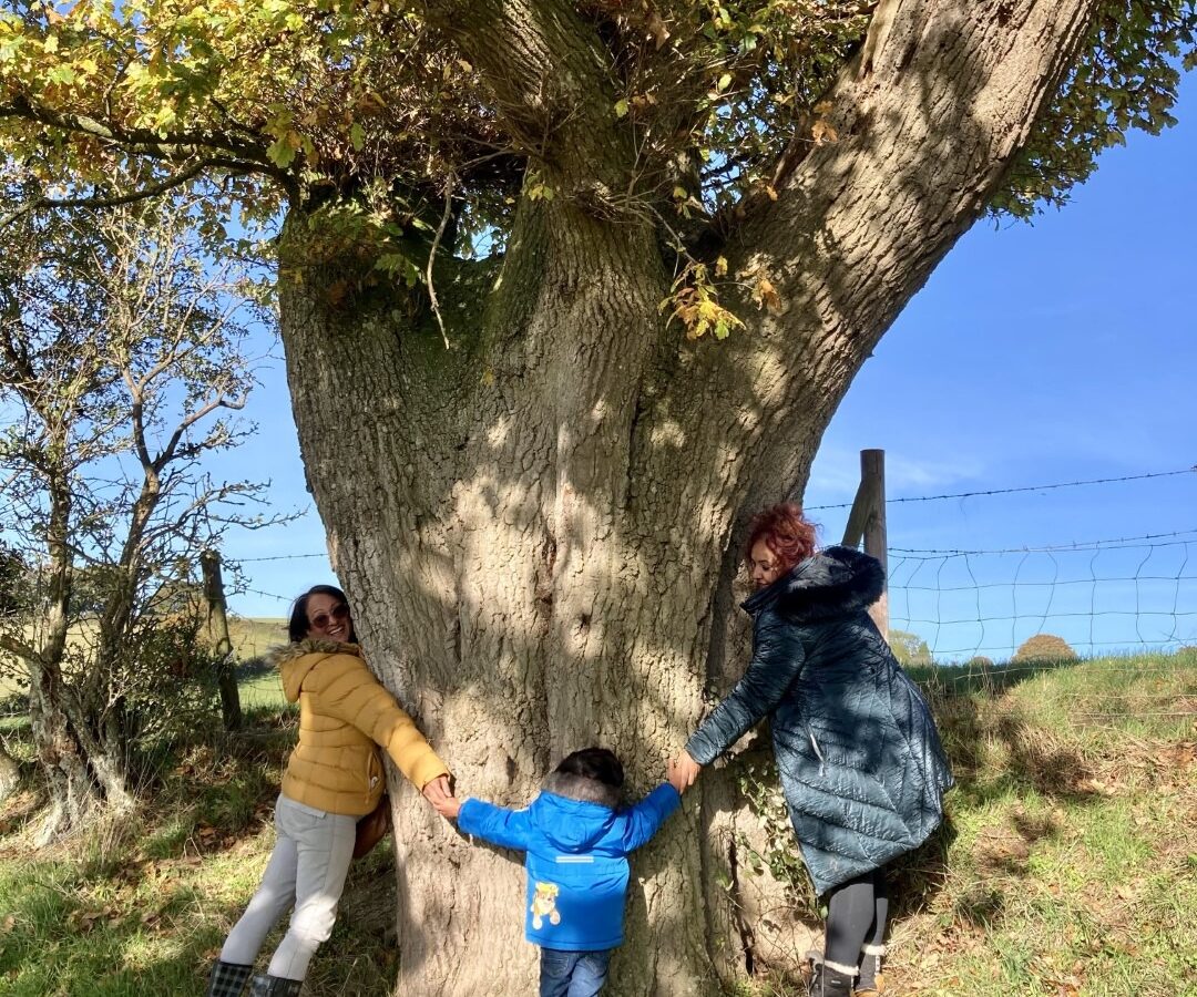 Two adults and a young child stand outside, holding hands around a large tree trunk in One Acre Wood. The adults wear warm jackets, one in yellow and the other in blue. The child wears a blue jacket and faces the tree. It is a sunny day with a clear blue sky and a bit of greenery around.
