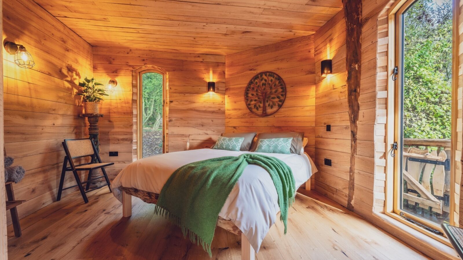 A cozy wooden cabin bedroom in One Acre Wood with a double bed, draped with a green blanket and white sheets. The room has wooden walls and floor, a small chair, and a leafy plant. Large windows let in natural light and offer a view of the surrounding trees.