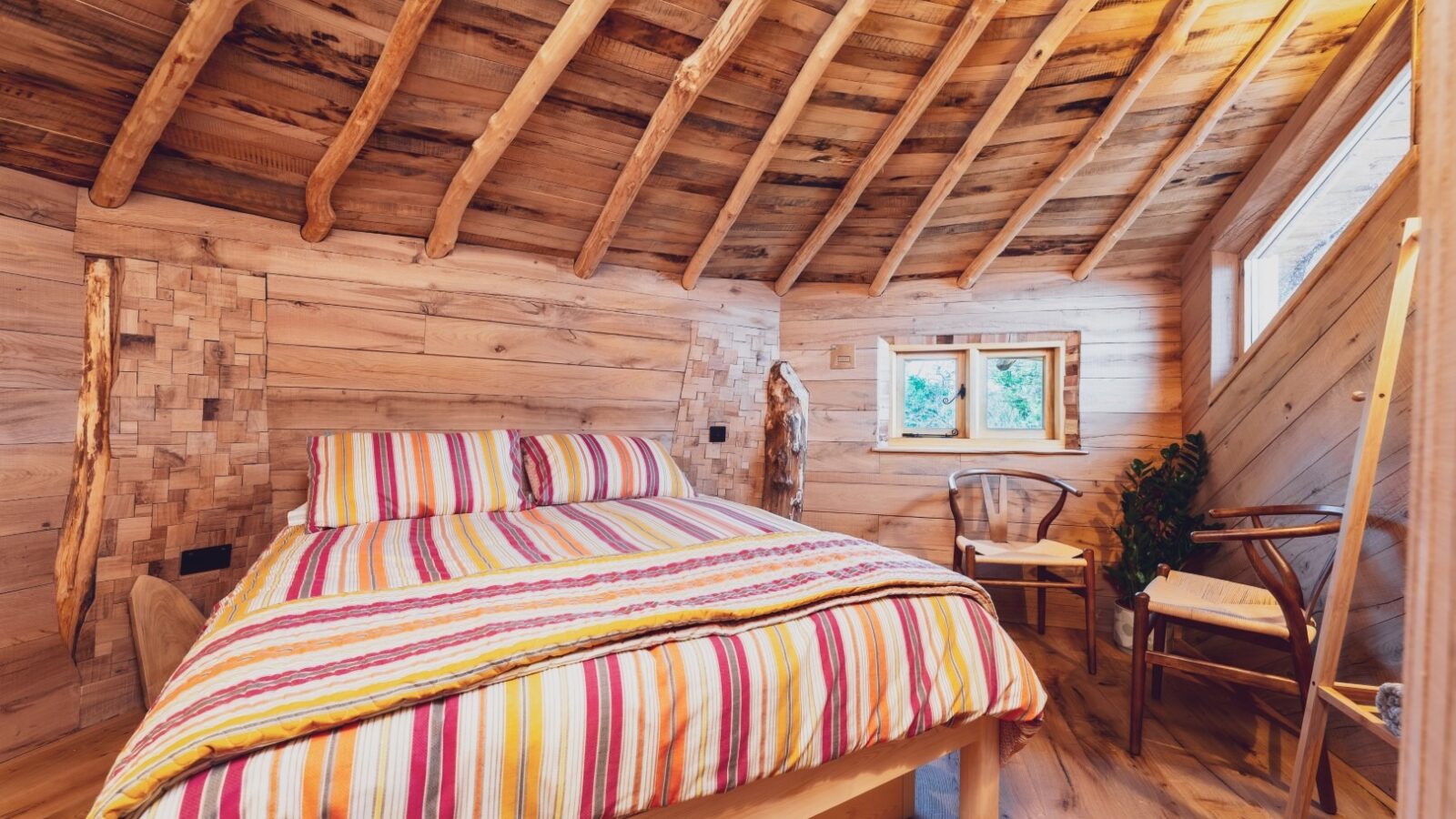 A cozy bedroom with a slanted wooden ceiling and walls reminiscent of One Acre Wood. The bed, with a colorful striped blanket and pillows, is positioned in the center. A small window with a view of greenery sits above a wooden nightstand. Two wooden chairs are placed near the window and door.