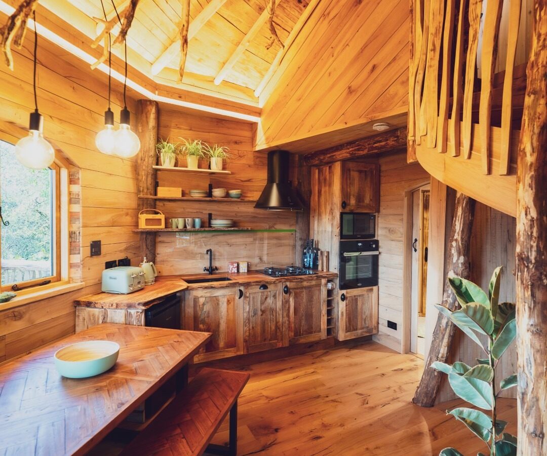 A cozy, rustic cabin interior in the heart of One Acre Wood features a wooden kitchen and dining area. The kitchen boasts wooden cabinets, black appliances, and potted plants. The dining table is flanked by bench seating. Warm lighting illuminates the space, highlighting natural wood beams and ceiling.