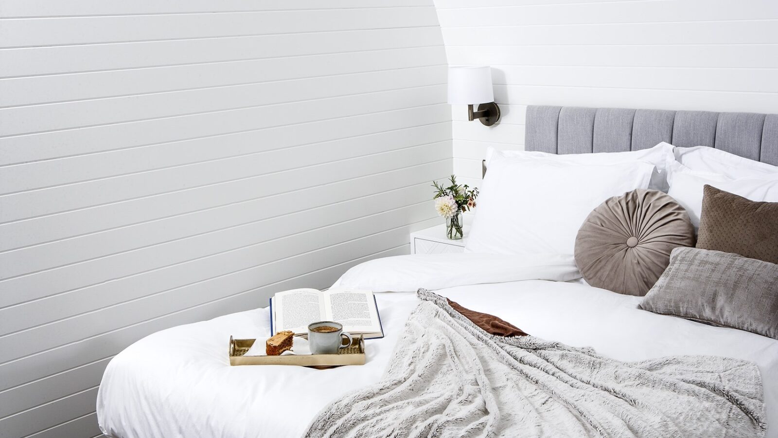 A cozy, bright bedroom with white walls and bedding. A gray upholstered headboard and assorted pillows add texture. On the bed are an open book, a tray with a mug and snack, and a beige knitted throw blanket. Inspired by glamping, a wall sconce and small vase of flowers adorn the bedside table.