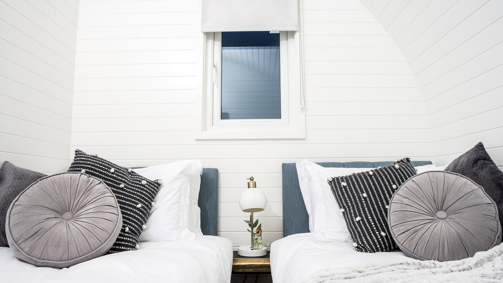 A cozy Pathgreen bedroom featuring two single beds side by side, each with white linens and decorative pillows. Between the beds is a small nightstand with a round lamp and a small plant. A window with a blind is on the wall above the nightstand, adding to the glamping-inspired ambiance.