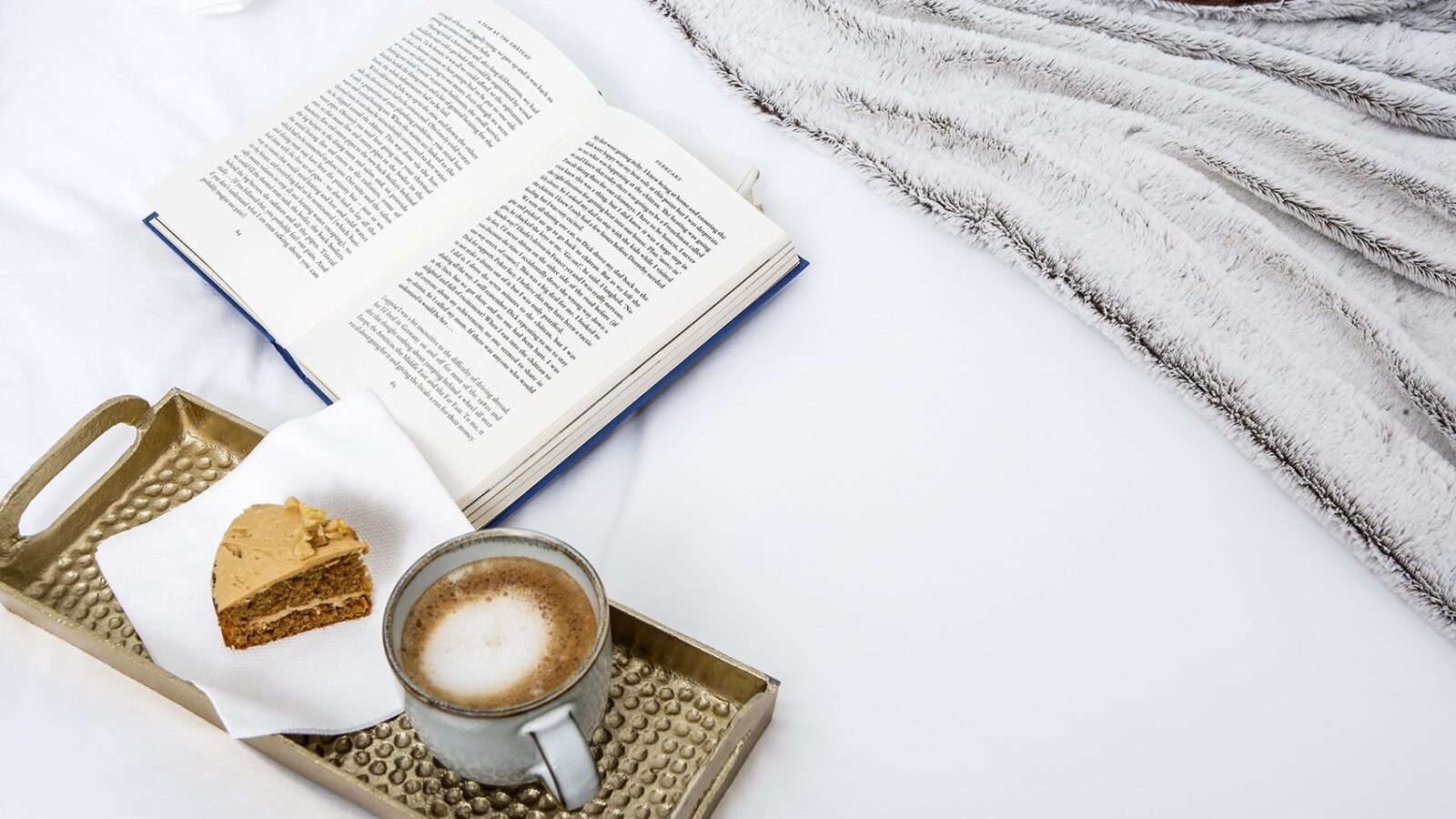 An open book rests on a white bedspread next to a cozy beige blanket. In front of the book, a tray holds a slice of cake on a napkin and a cup of frothy coffee, invoking the relaxed and comfortable vibe of Pathgreen Glamping.