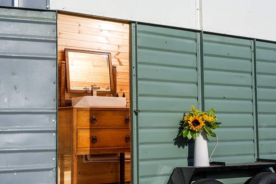 A small, wooden-paneled bathroom with a vintage-style wooden vanity and a mirror is visible through the open door of the glamping structure. A white enamel pitcher filled with yellow flowers sits on an exterior ledge next to the door, adding a touch of Pathgreen charm.