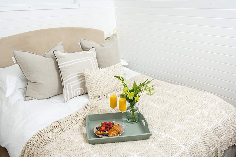 A cozy bedroom in Pathgreen features a neatly made bed with a beige knitted blanket, patterned pillows, and two glasses of orange juice on a tray. The tray also holds a croissant and a small bowl of assorted berries, next to a vase with green and white flowers—perfect for glamping indoors.