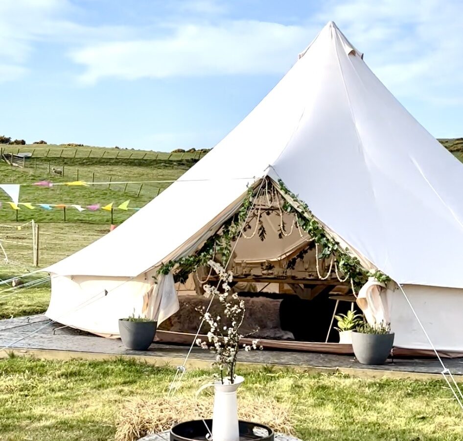 A large white Pathgreen canvas tent erected outdoors on a grassy field, decorated with greenery and fairy lights around the entrance. Inside, furniture and decor are slightly visible. In the foreground, a wooden spool table holds a vase with white flowers and a small tray.