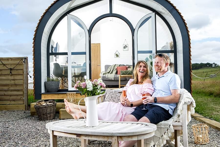 A couple relaxes on an outdoor sofa, holding drinks and smiling, in front of a cozy, arched tiny house with large windows at Pathgreen Glamping. A flower vase sits on a nearby table. The scene is set in an open, rural area on a sunny day.