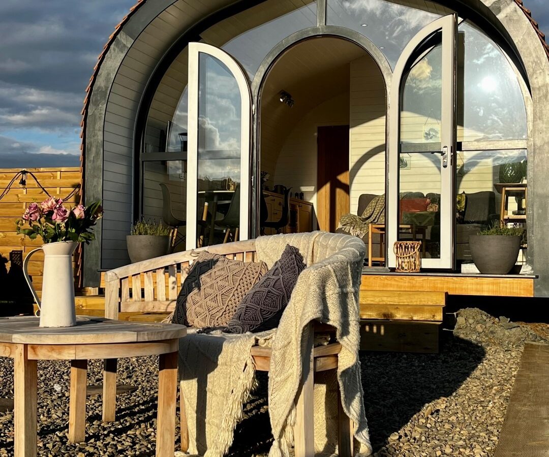 A cozy wooden cabin with large arched glass doors open, revealing a warmly lit interior. Outside, a small wooden table with a pitcher of flowers and a bench draped with a knitted blanket and cushions sit on gravel, epitomizing glamping at its finest under the partly cloudy sky.